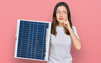 Young beautiful girl holding photovoltaic solar panel serious face thinking about question with...