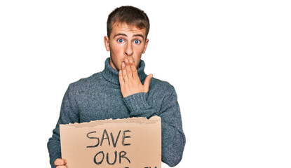 Young blond man holding save our democracy protest banner covering mouth with hand, shocked and...