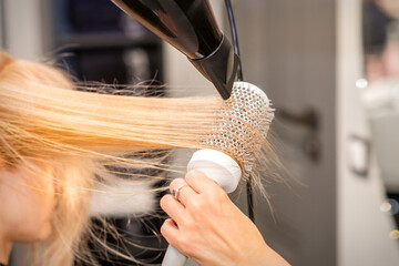 Drying straight blond hair with black hairdryer and white round brush in hairdresser salon, close up