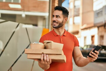 Young hispanic man using smartphone holding take away food at the city.