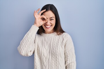 Young brunette woman standing over blue background doing ok gesture with hand smiling, eye looking through fingers with happy face.