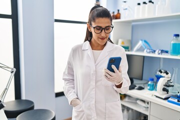 Obraz na płótnie Canvas Young hispanic woman wearing scientist uniform using smartphone at laboratory