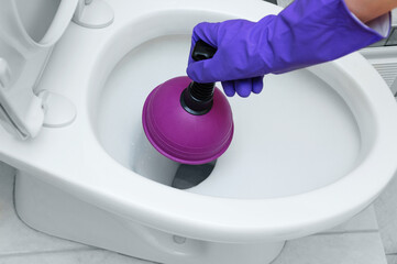 A hand in a protective glove cleans blockage in the toilet bowl with a plunger.