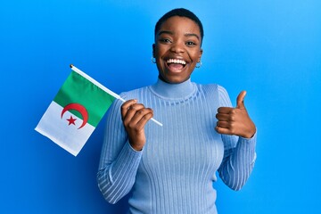 Young african american woman holding algeria flag smiling happy and positive, thumb up doing excellent and approval sign - obrazy, fototapety, plakaty