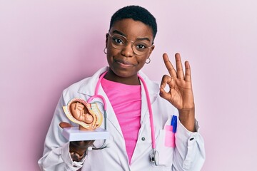 Young african american doctor woman holding anatomical model of female uterus with fetus doing ok...