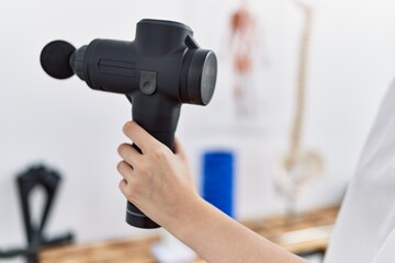 Young caucasian woman wearing physio therapist uniform using percussion pistol at clinic