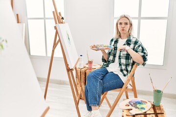 Young artist woman painting on canvas at art studio puffing cheeks with funny face. mouth inflated with air, crazy expression.