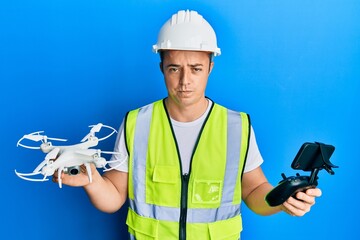 Handsome young man wearing safety hard using drone and control skeptic and nervous, frowning upset...