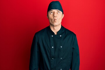 Handsome young man wearing professional cook uniform and hat making fish face with lips, crazy and comical gesture. funny expression.