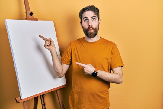 Caucasian man with beard pointing to painter easel stand making fish face with mouth and squinting eyes, crazy and comical.