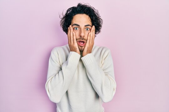Handsome hispanic man wearing casual white sweater afraid and shocked, surprise and amazed expression with hands on face