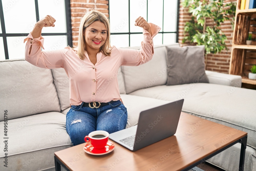 Sticker Young hispanic woman using laptop sitting on the sofa at home showing arms muscles smiling proud. fitness concept.