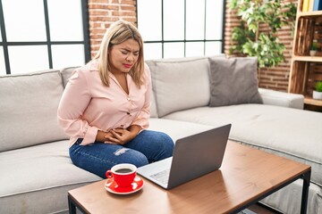 Young hispanic woman using laptop sitting on the sofa at home with hand on stomach because nausea, painful disease feeling unwell. ache concept.