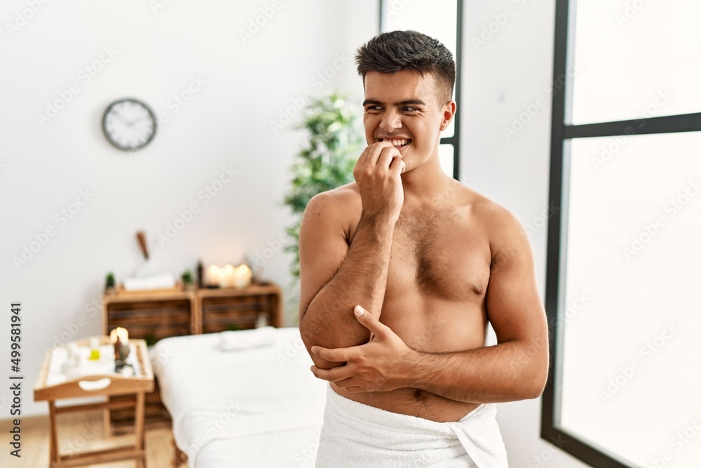 Sticker Young hispanic man standing shirtless at spa center looking stressed and nervous with hands on mouth biting nails. anxiety problem.