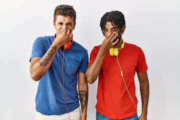 Young hispanic brothers standing over isolated background wearing headphones smelling something stinky and disgusting, intolerable smell, holding breath with fingers on nose. bad smell