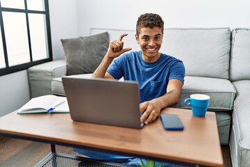Young handsome hispanic man using laptop sitting on the floor smiling and confident gesturing with hand doing small size sign with fingers looking and the camera. measure concept.