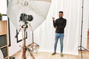 Young hispanic man with beard posing as model at photography studio with a big smile on face, pointing with hand and finger to the side looking at the camera.
