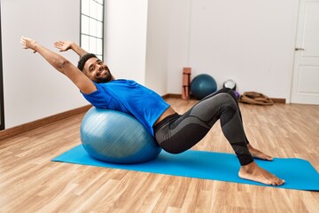 Fototapeta na wymiar Handsome hispanic man doing exercise and stretching on yoga mat, practicing flexibility with pilates ball at the gym