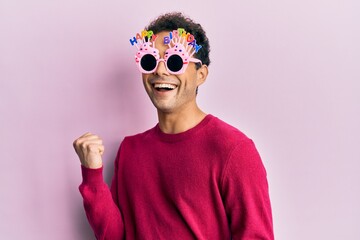 Handsome hispanic man wearing funny happy birthday glasses celebrating surprised and amazed for...