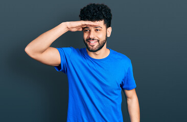 Young arab man with beard wearing casual blue t shirt very happy and smiling looking far away with hand over head. searching concept.