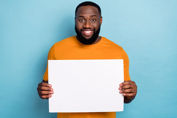 Photo of funny sweet guy dressed orange t-shirt holding white paper sheet empty space isolated blue...