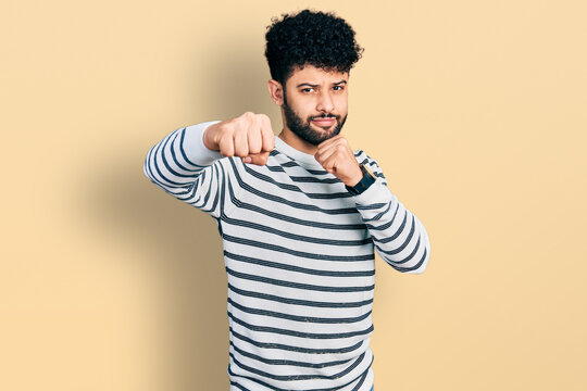 Young arab man with beard wearing casual striped sweater punching fist to fight, aggressive and angry attack, threat and violence