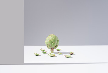 Una alcachofa cruda en pie con hojas que caen sobre una mesa blanca y fondo gris. Minimalista	