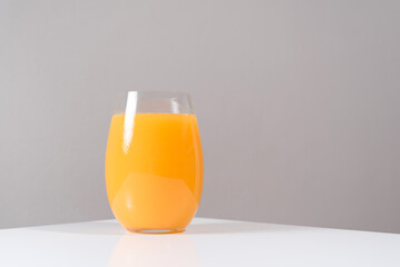 Un vaso de zumo de naranja exprimido en un vaso de cristal con naranja sobre una mesa blanca con un fondo gris	 - obrazy, fototapety, plakaty