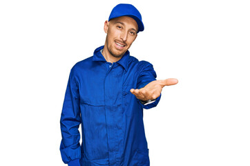 Bald man with beard wearing builder jumpsuit uniform smiling cheerful offering palm hand giving assistance and acceptance.