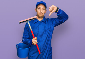 Bald man with beard wearing glass cleaner uniform and squeegee with angry face, negative sign showing dislike with thumbs down, rejection concept