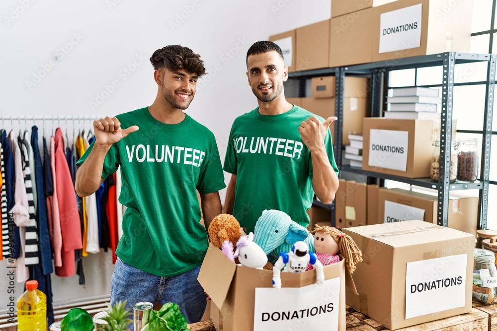 Canvas Prints Young gay couple wearing volunteer t shirt at donations stand pointing to the back behind with hand and thumbs up, smiling confident