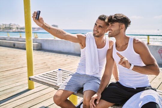 Two hispanic men sporty couple make selfie by the smartphone sitting on bench at seaside