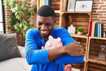 Young african american man hugging piggy bank sitting on sofa at home