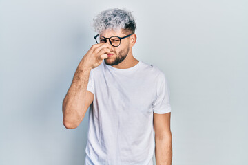 Young hispanic man with modern dyed hair wearing white t shirt and glasses smelling something stinky and disgusting, intolerable smell, holding breath with fingers on nose. bad smell