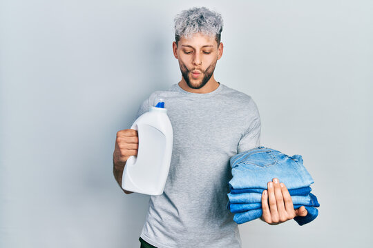 Young hispanic man with modern dyed hair holding jeans for laundry and detergent bottle making fish face with mouth and squinting eyes, crazy and comical.