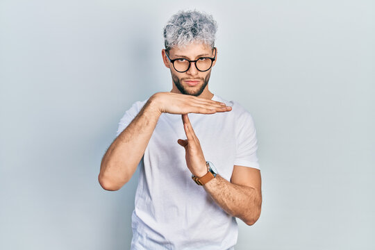 Young hispanic man with modern dyed hair wearing white t shirt and glasses doing time out gesture with hands, frustrated and serious face