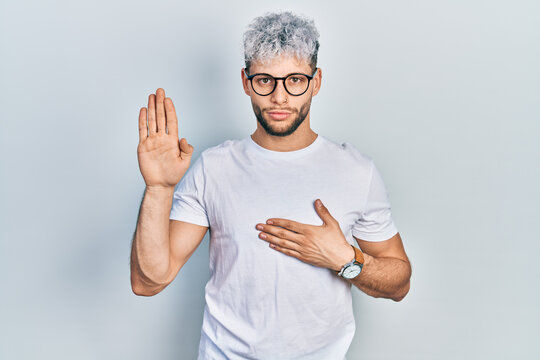 Young hispanic man with modern dyed hair wearing white t shirt and glasses swearing with hand on chest and open palm, making a loyalty promise oath