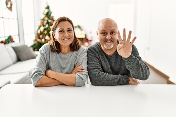 Middle age hispanic couple sitting on the table by christmas tree showing and pointing up with fingers number four while smiling confident and happy.