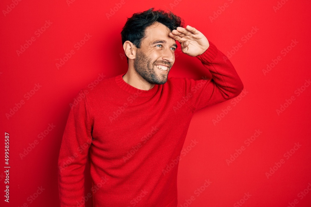 Poster Handsome man with beard wearing casual red sweater very happy and smiling looking far away with hand over head. searching concept.