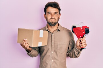 Handsome man with beard holding packing tape holding cardboard puffing cheeks with funny face....