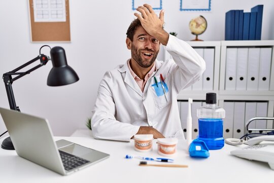 Young hispanic dentist man working at medical clinic surprised with hand on head for mistake, remember error. forgot, bad memory concept.