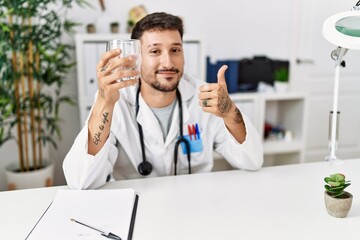 Young doctor holding glass of water smiling happy and positive, thumb up doing excellent and approval sign