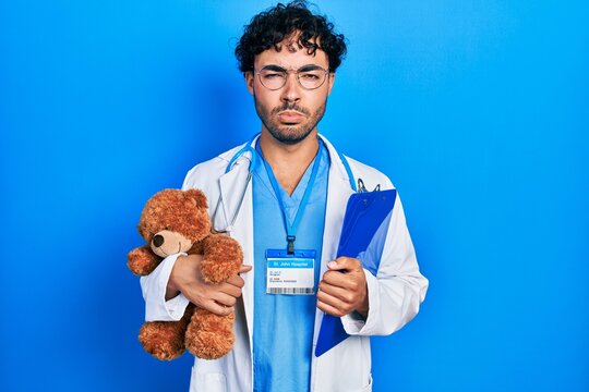 Young hispanic man wearing doctor uniform holding teddy bear and clipboard skeptic and nervous, frowning upset because of problem. negative person.
