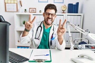 Young man with beard wearing doctor uniform and stethoscope at the clinic smiling with tongue out...