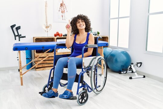 Young middle eastern woman sitting on wheelchair at physiotherapy clinic pointing finger up with successful idea. exited and happy. number one.