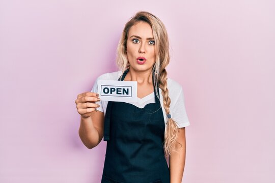 Beautiful young blonde woman wearing waitress apron holding open banner scared and amazed with open mouth for surprise, disbelief face
