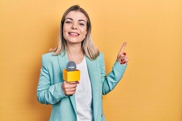 Beautiful caucasian woman holding reporter microphone smiling happy pointing with hand and finger to the side