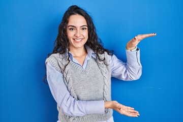 Young brunette woman standing over blue background gesturing with hands showing big and large size sign, measure symbol. smiling looking at the camera. measuring concept.