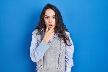 Obraz premium Young brunette woman standing over blue background looking fascinated with disbelief, surprise and amazed expression with hands on chin