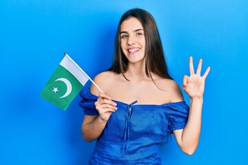 Young brunette teenager holding pakistan flag doing ok sign with fingers, smiling friendly gesturing excellent symbol